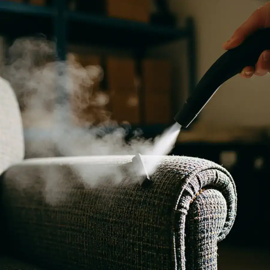 Close-up of a steam cleaner nozzle releasing a jet of steam into the seam of a vintage upholstered armchair.