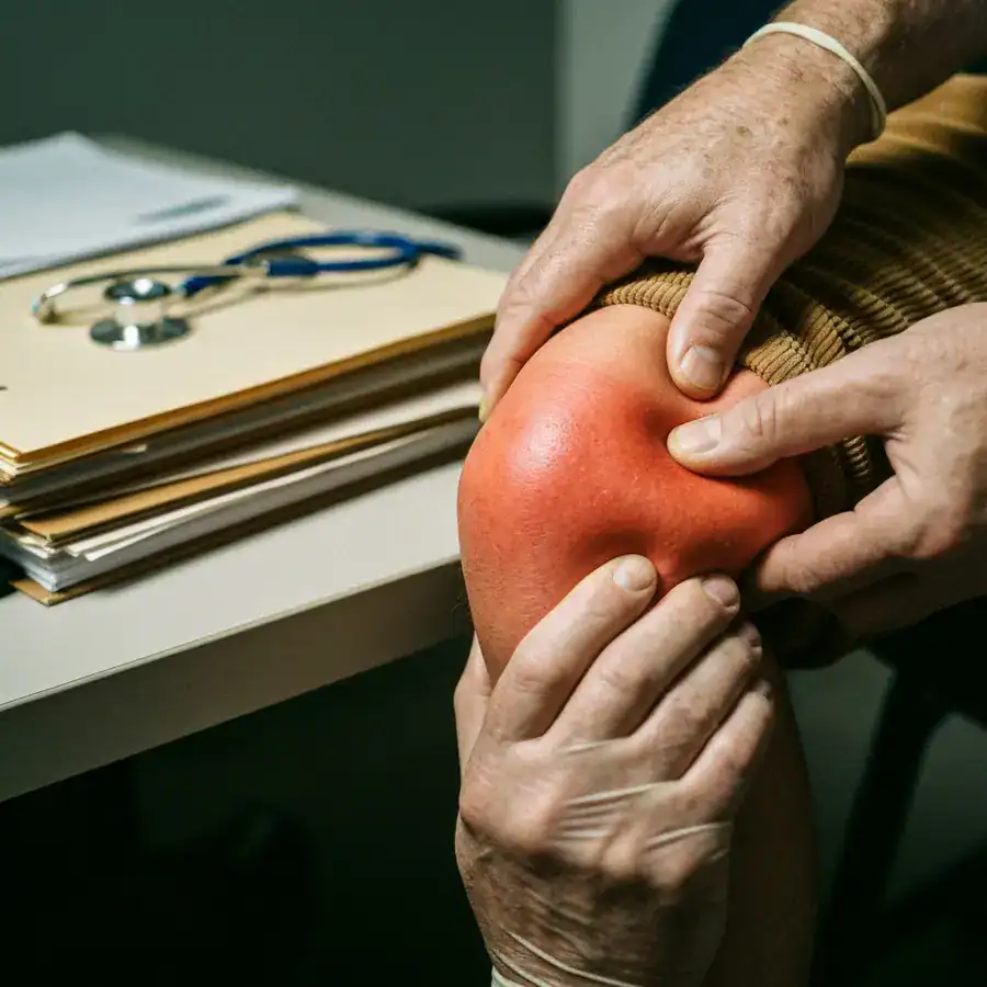 Gloved hands palpating a swollen, red knee joint on a patient in a dimly lit medical clinic.