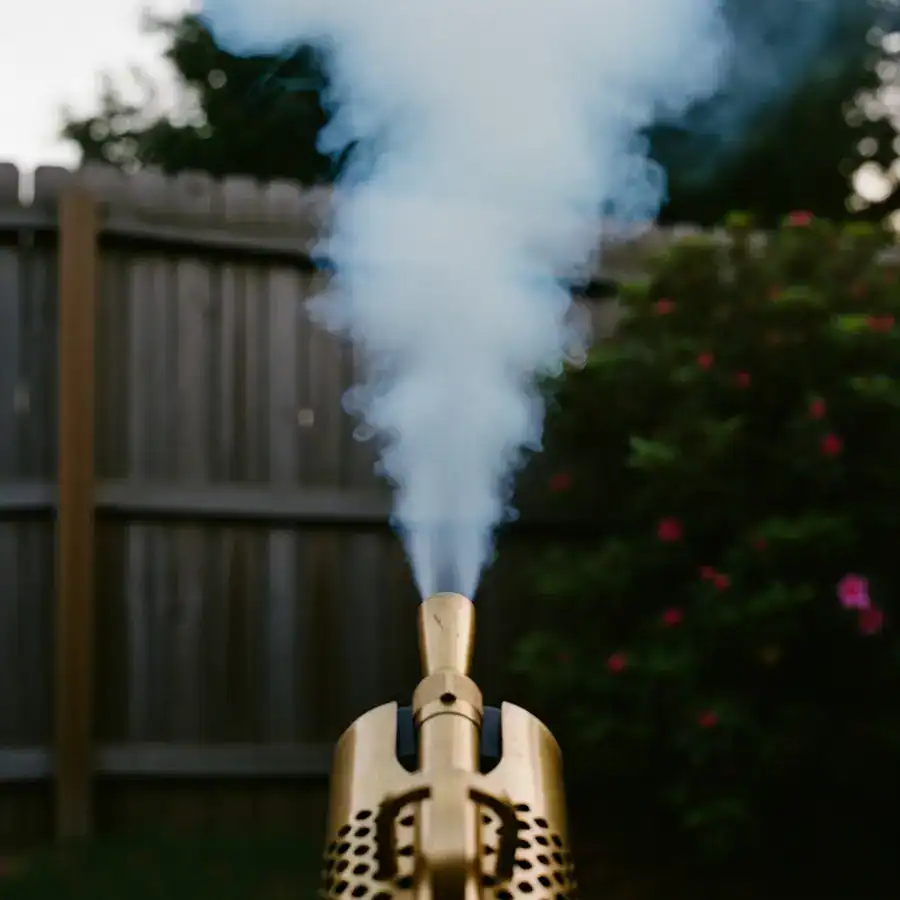 A propane thermal fogger nozzle emits a thick white fog that drifts upwards and away from bushes in a backyard at dusk.